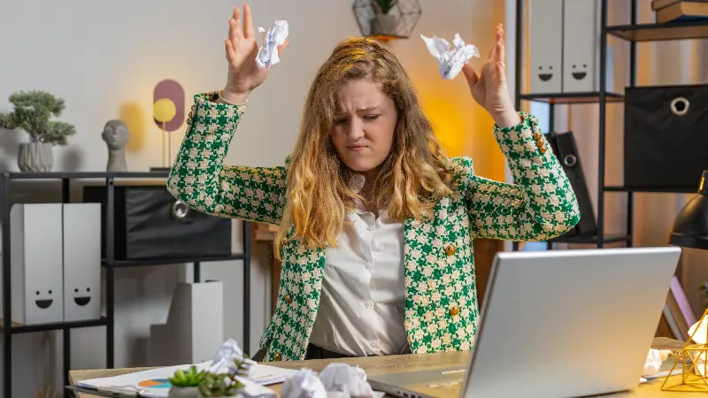 Žena za radnim stolom baca zgužvane papire u vazduh, pokazujući frustraciju i stres tokom rada na laptopu.