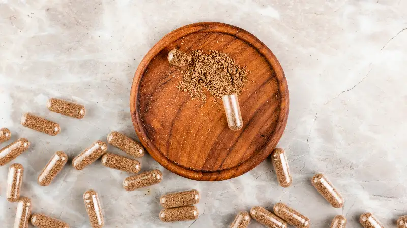 Kapsule s biljnim prahom rasute po površini, uz drvenu posudicu s prosutim sadržajem kapsule.