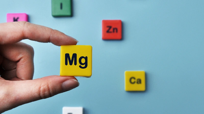 Ruka drži simbol Mg (magnezijum) okružen drugim mineralima poput cinka, kalcijuma i gvožđa, što ukazuje na važnu ulogu magnezijuma u sprečavanju grčeva u nogama i pravilnom funkcionisanju organizma