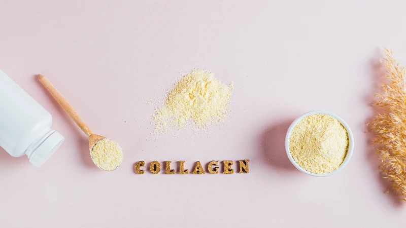 Bočica i drvena kašika sa kolagen prahom, rasuti kolagen i činija sa prahom na svetloj pozadini, uz dekorativnu suvu travu.