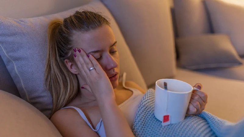 Umorna žena leži na kauču sa šoljom čaja, drži se za glavu i pokazuje simptome prehlade ili glavobolje u kućnom ambijentu