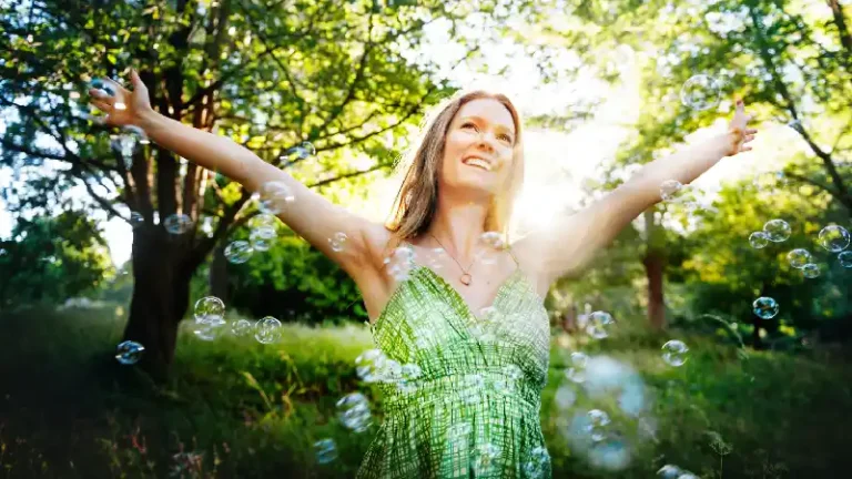 Žena u zelenoj haljini sa podignutim rukama uživa u prirodi dok oko nje lete mehurići, osunčana šumska pozadina u toplom letnjem ambijentu