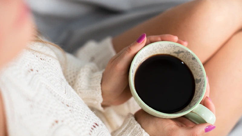 Osoba drži šolju crne kafe u opuštenoj atmosferi, simbol jutarnje rutine, energije i uživanja u toplom napitku, i da kafa utiče na menstruaciju i hormone
