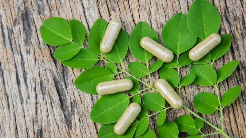 Kapsule moringa ekstrakta položene na sveže listove moringe na drvenoj podlozi, prikaz dodatka ishrani Moringa Ekstrakt za prirodnu podršku energiji, vitalnosti i opštem zdravlju.